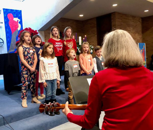 A childrens choir singing and their director playing guitar.